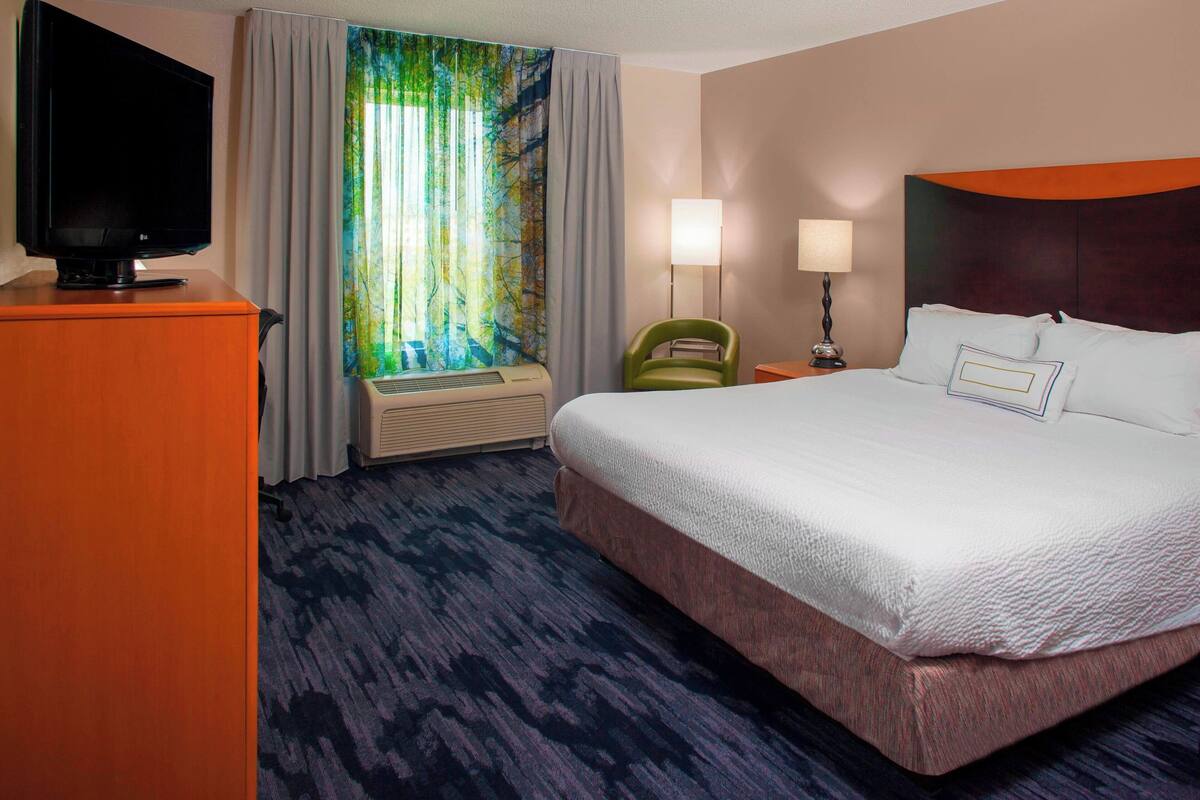 A hotel room with a large bed, a television on a wooden cabinet, a window with curtains, and a nightstand with a lamp.