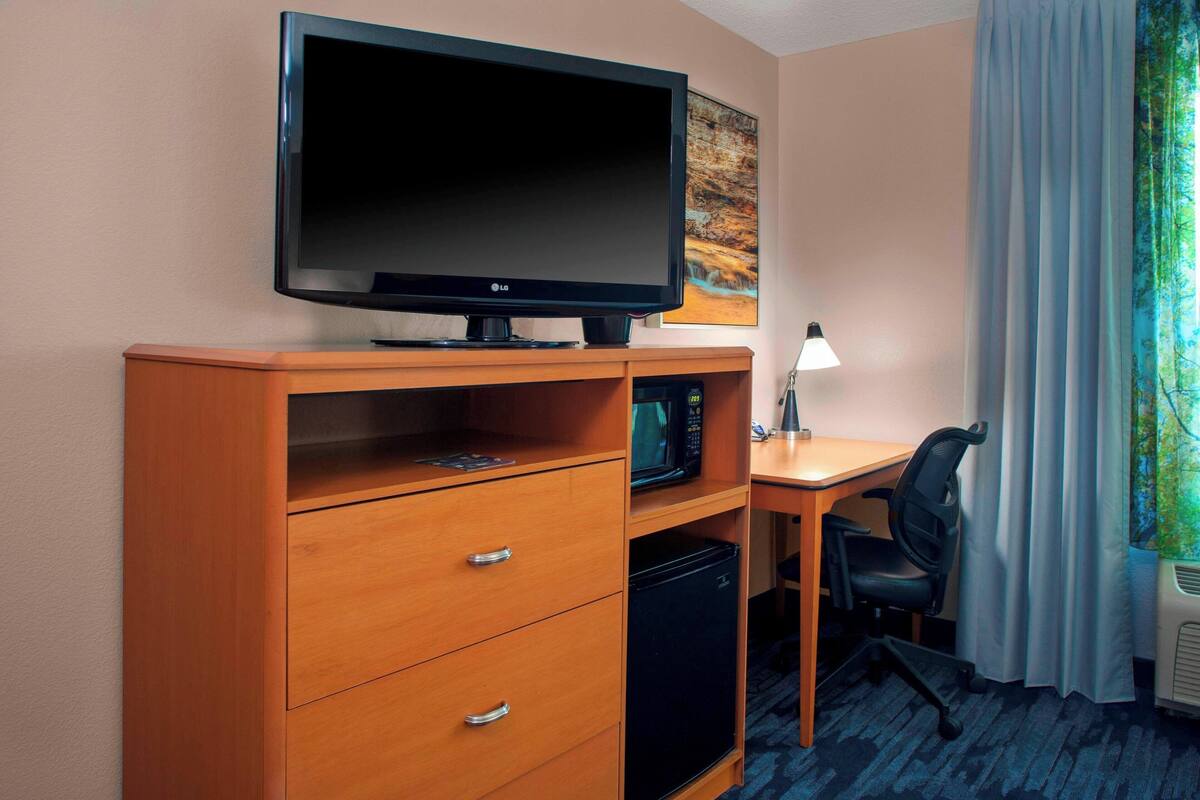 A hotel room with a flat-screen TV, a microwave, a mini fridge, a desk with a chair, and a window with curtains.