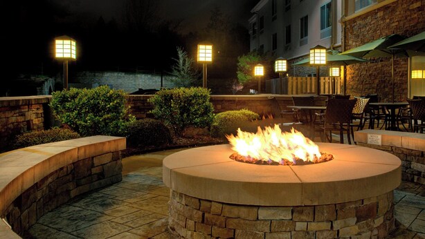 A circular stone fire pit with flames, surrounded by stone walls and benches, under outdoor lighting. - Fairfield Inn & Suites by Marriott Montgomery EastChase Pkwy (Montgomery)