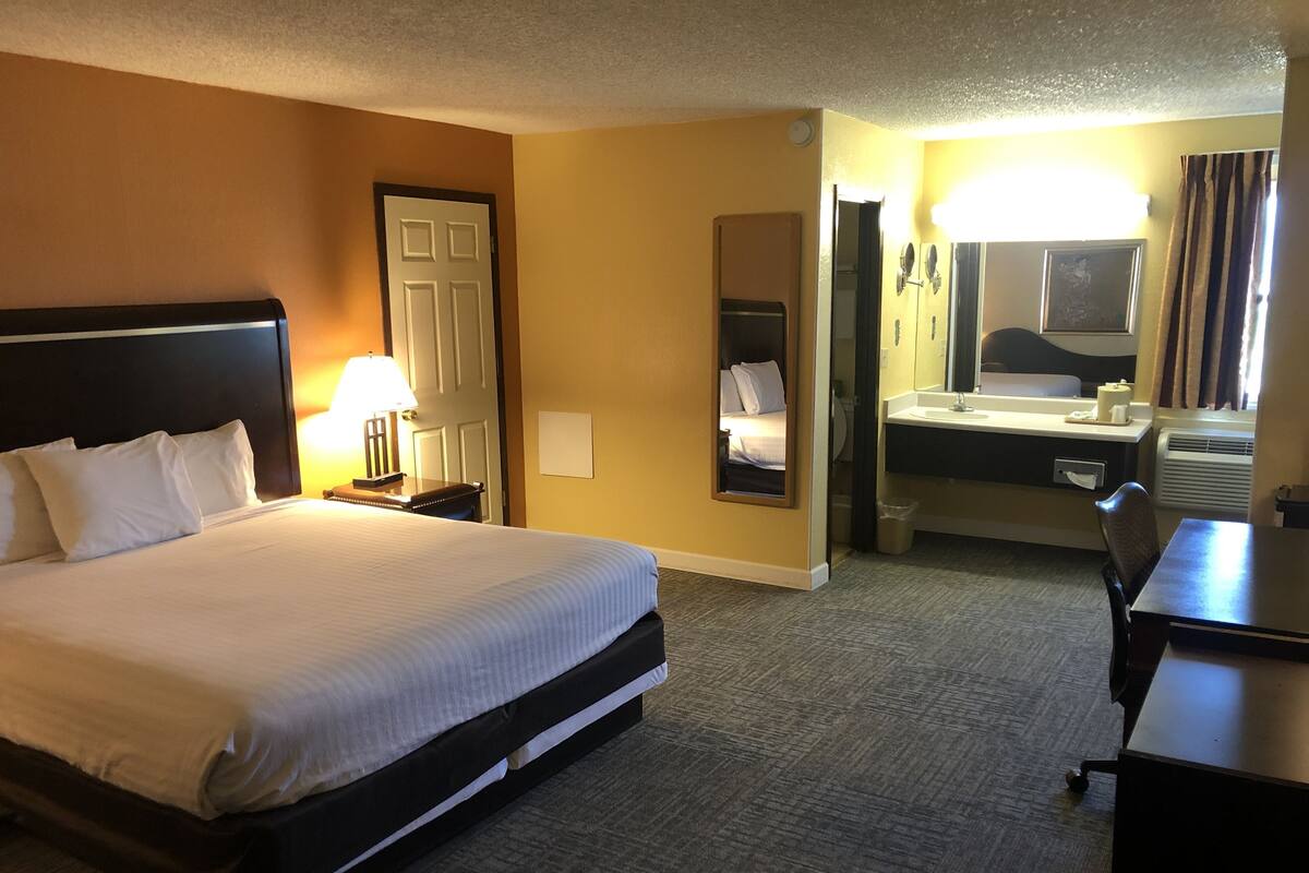 A hotel room with a large bed, a desk, a chair, a desk lamp, a mirror, a bathroom with a sink and toilet, and a door.