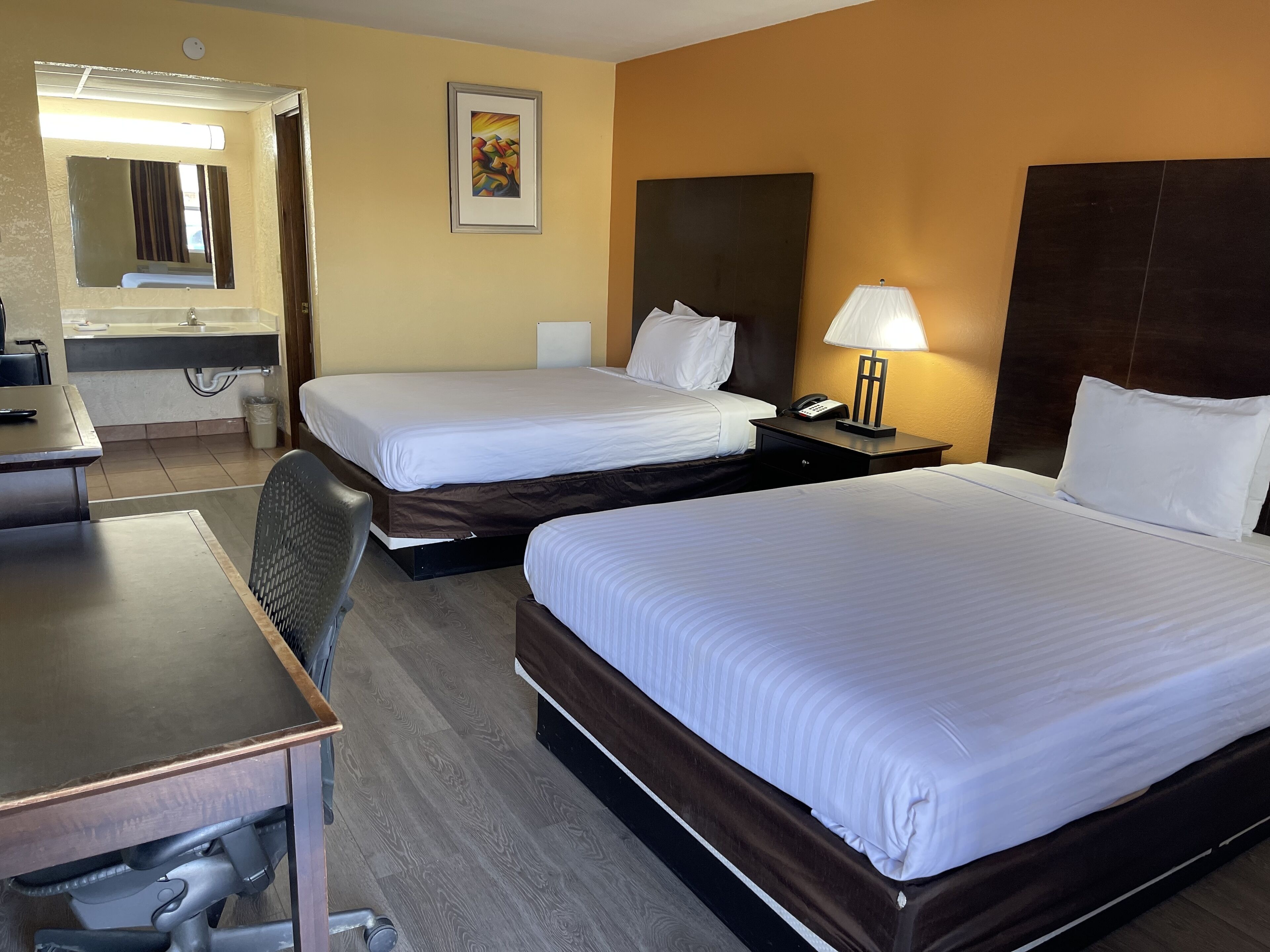 A hotel room with two beds, a desk, a chair, a lamp, and a painting on the wall.