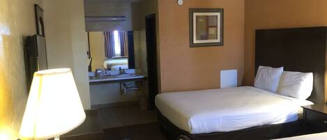 Habitación de hotel con dos camas, un escritorio con lámpara, baño visible a través de una puerta abierta y un cuadro en la pared.