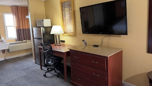 A hotel room with a desk, chair, TV, refrigerator, and air conditioning unit. - Nendels Inn & Suites (Dodge City)