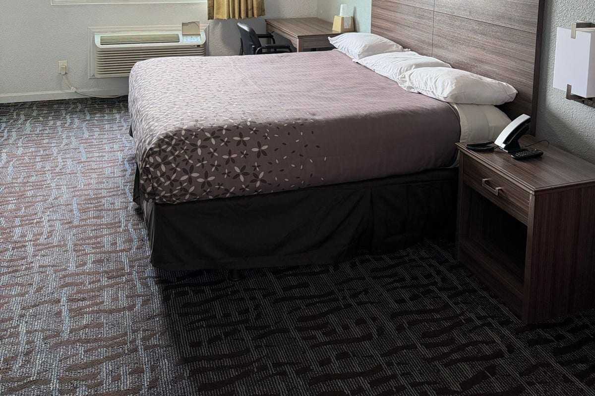 Una habitación de hotel con cama, mesita de noche, lámpara y ventana con cortinas.