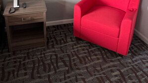 A hotel room with a red armchair, a small wooden nightstand with a phone and remote, and a wall-mounted lamp. - Regency Inn And Suites Nashville (Nashville)