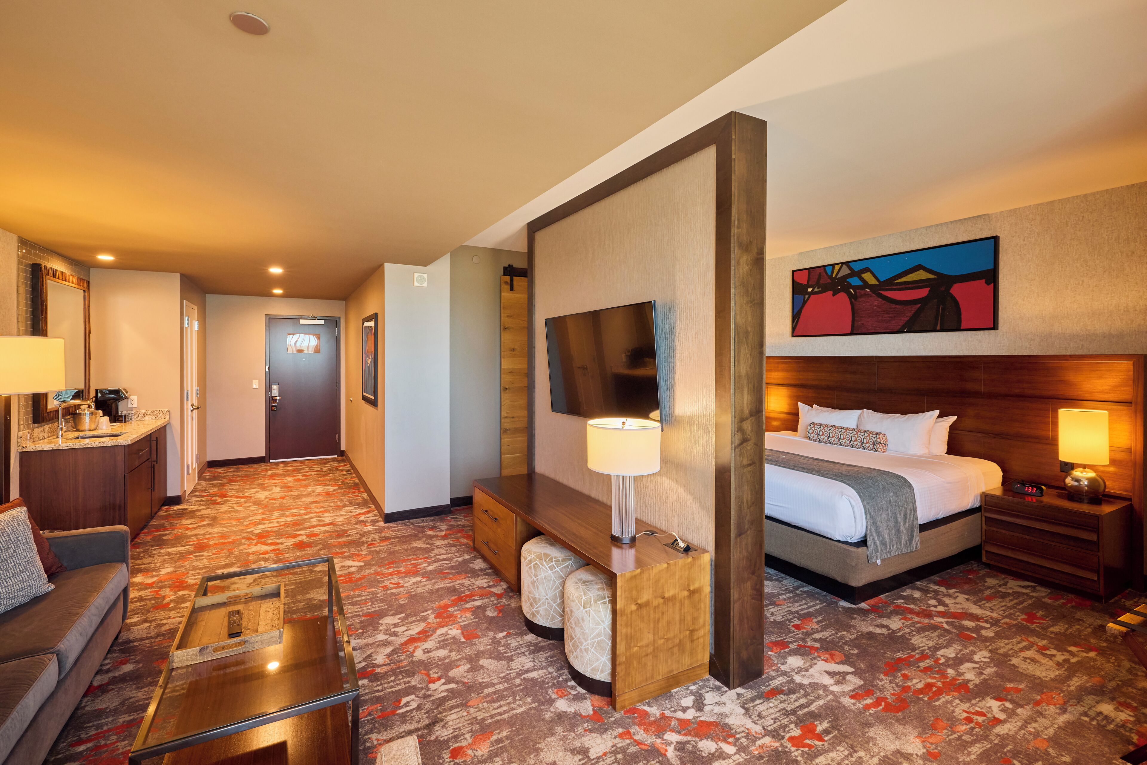 A hotel room with a bed, bedside table, lamp, TV, and a painting on the wall.