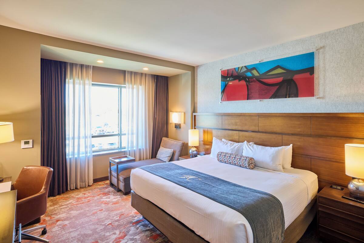 Habitación de hotel con una cama grande, un escritorio, una silla, un ventanal con cortinas, un cuadro en la pared y una lámpara en la mesita de noche.