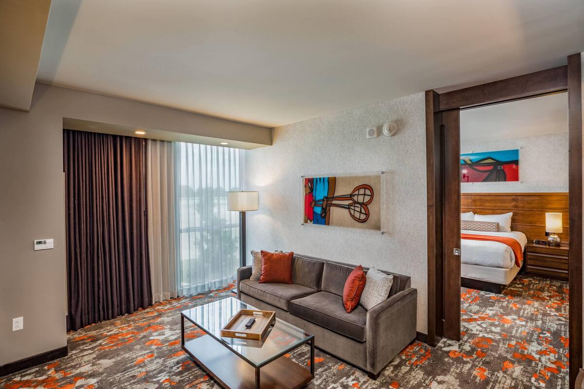 Habitación de hotel con sofá, mesa de centro, lámpara, cuadro y cama con mesita de noche.
