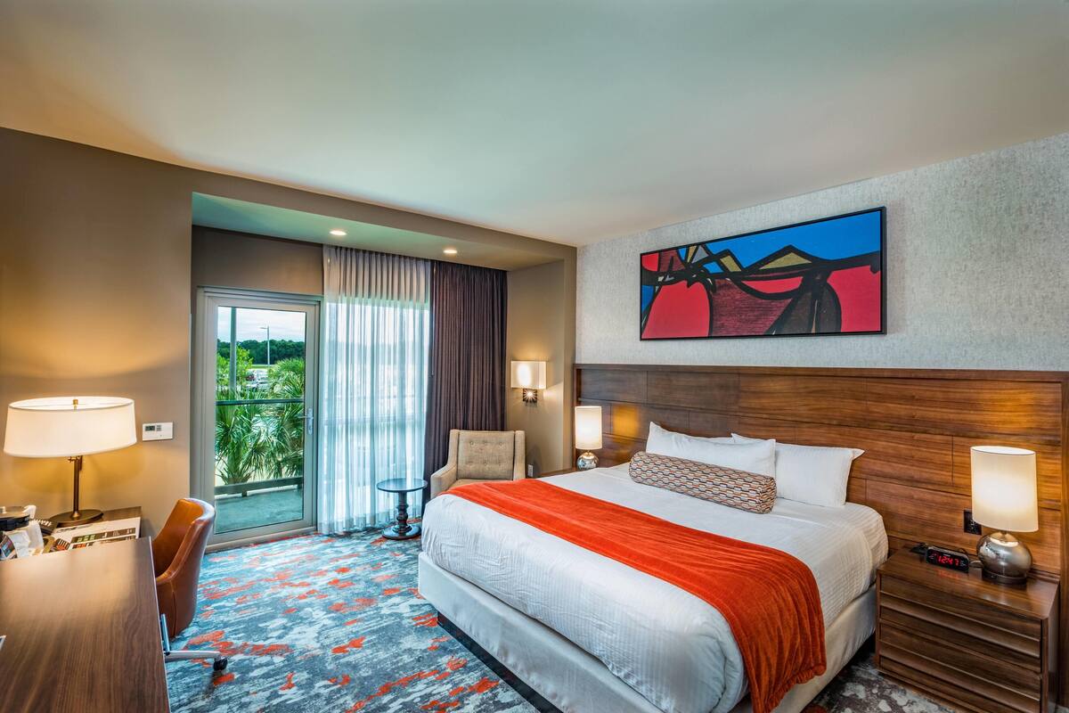Habitación de hotel con una cama grande, un escritorio, una silla, una mesita de noche y un cuadro en la pared.
