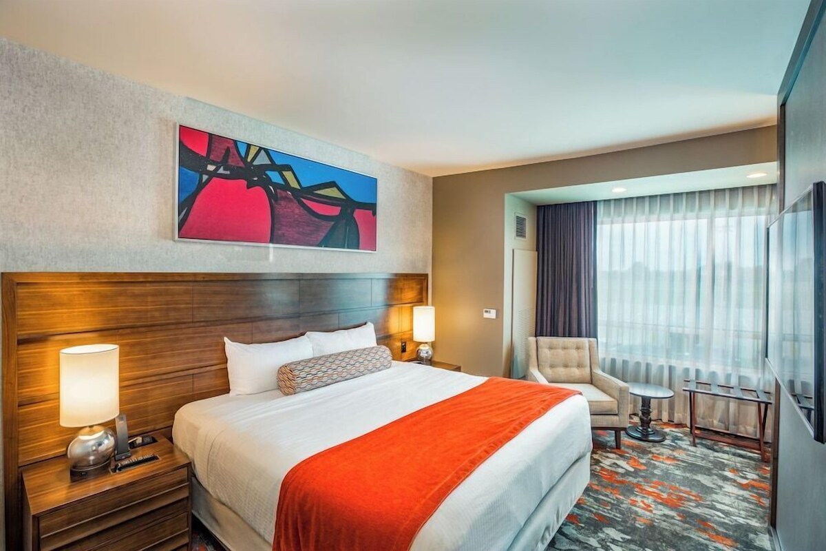 A hotel room with a large bed, two bedside lamps, a chair, a small table, a painting on the wall, and a view of the outdoors through the window.