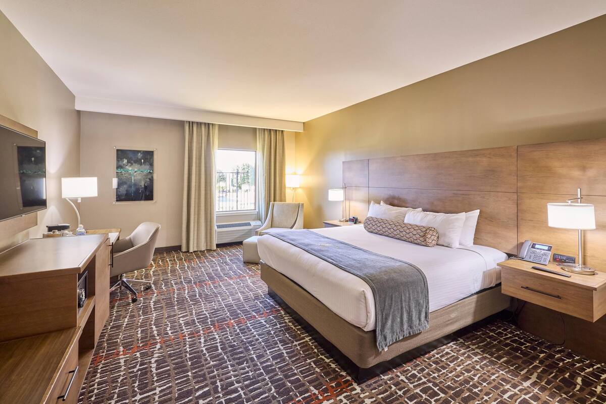 Habitación de hotel con cama, escritorio, silla, mesita de noche, lámpara y una obra de arte.