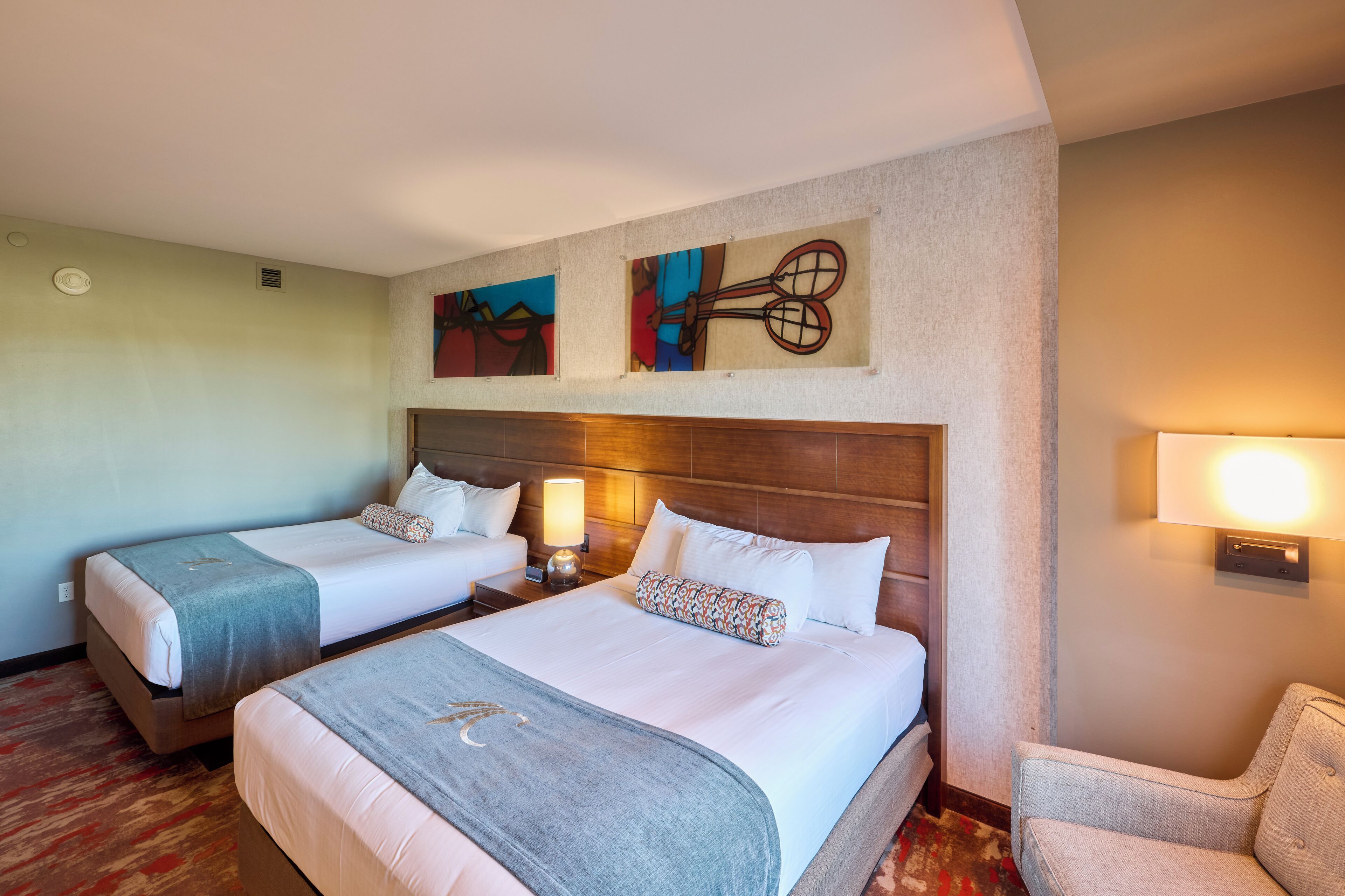 Una habitación de hotel con dos camas, una mesita de noche con lámpara, una silla y una obra de arte en la pared.