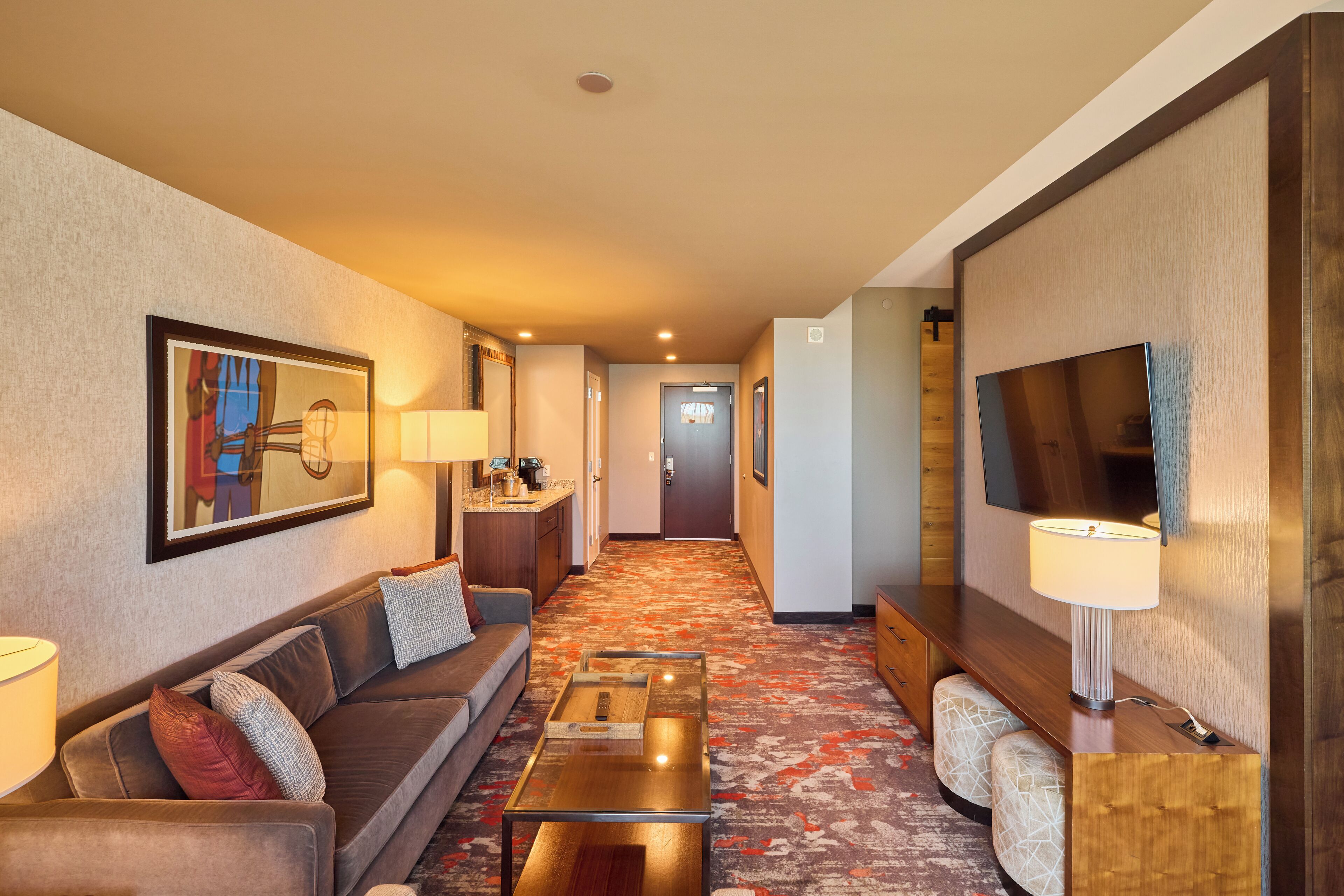 A hotel room with a sofa, a coffee table, a TV, a lamp, and a hallway leading to a door.