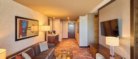 A hotel room with a sofa, a coffee table, a TV, a lamp, and a hallway leading to a door.