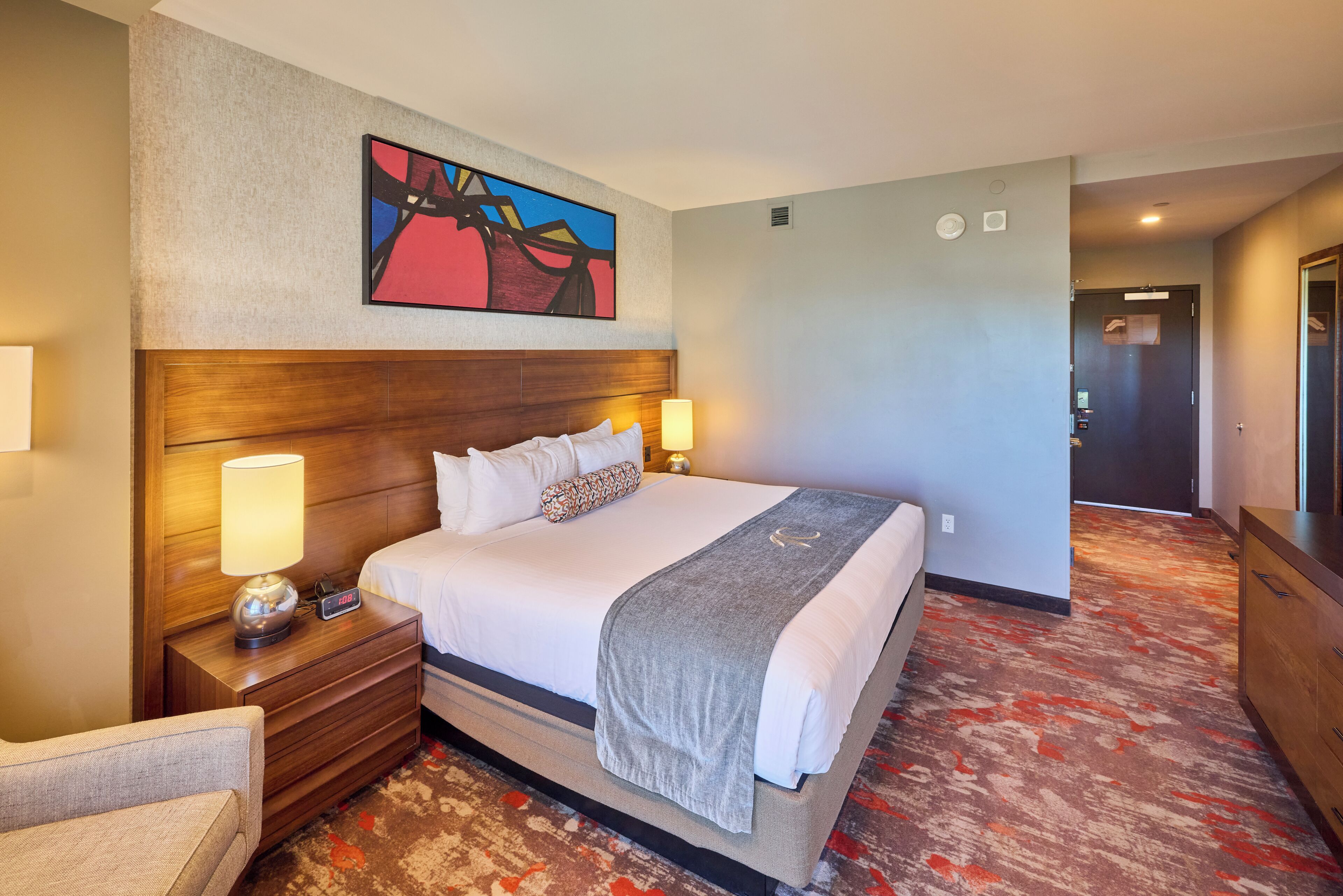 Une chambre d’hôtel avec un grand lit, des lampes de chevet, une tête de lit, un tableau au mur et un sol recouvert de moquette.