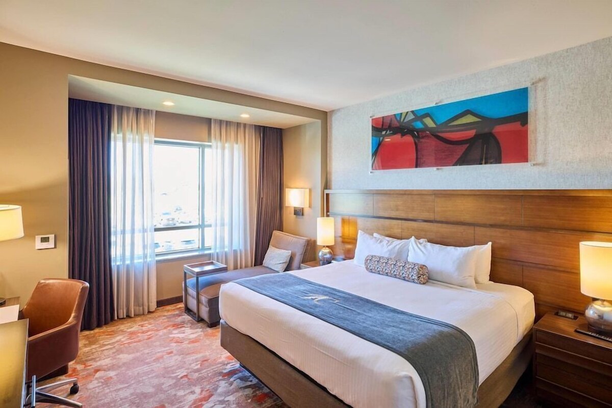 A hotel room with a large bed, a desk, a chair, a window with curtains, and a painting on the wall.