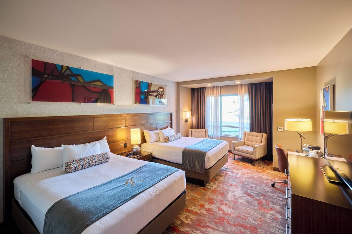 Habitación de hotel con dos camas, un escritorio, una silla y obras de arte en las paredes.