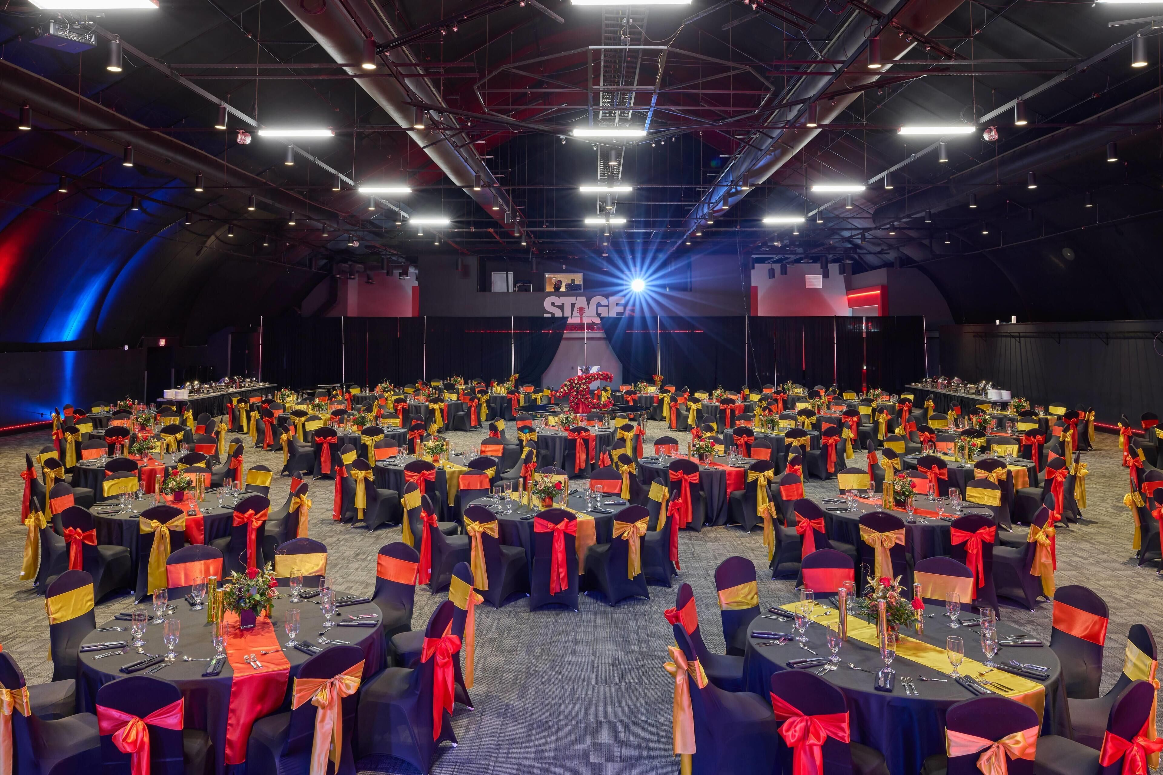 A large indoor event space with rows of round tables, each set with chairs and table settings, and a stage area in the background.