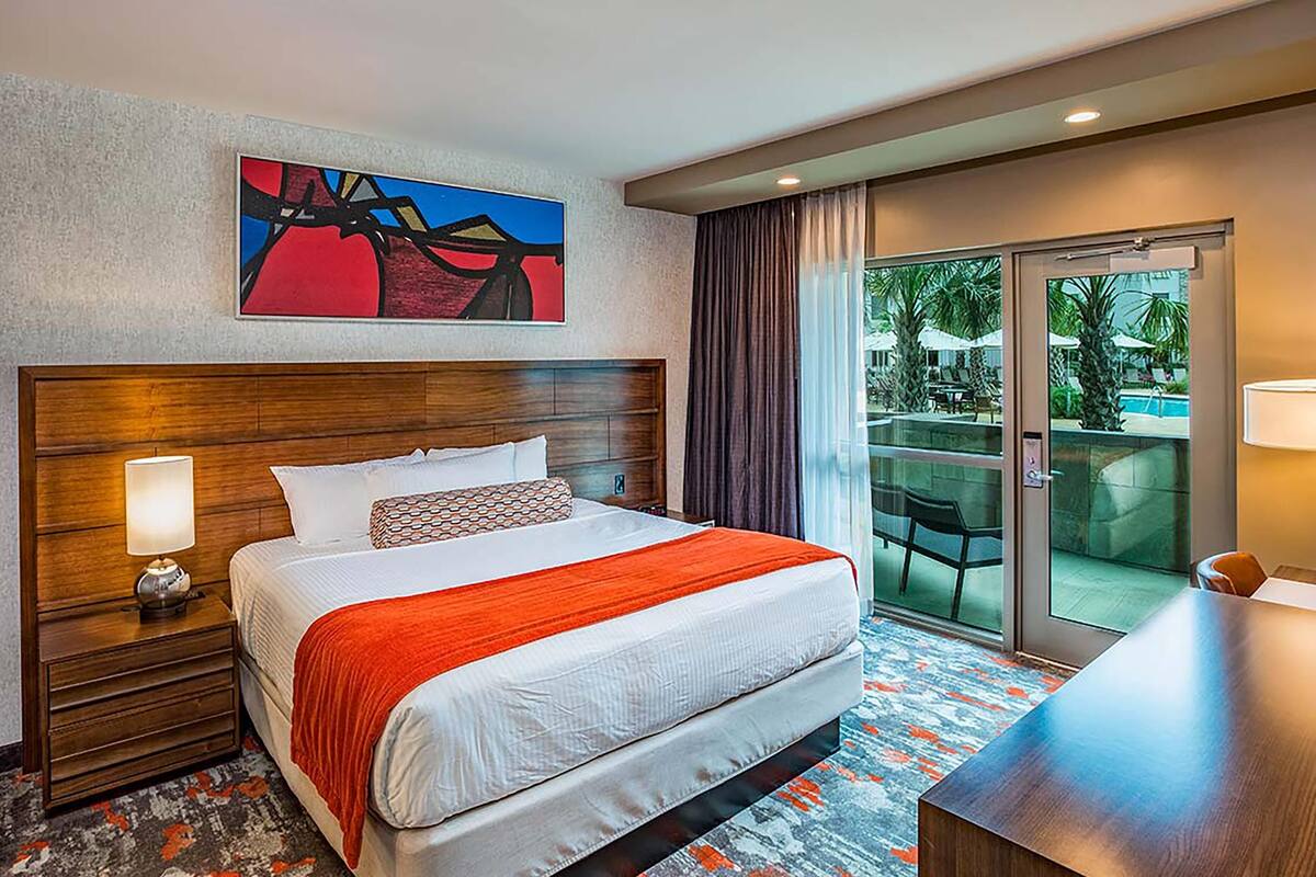 Habitación de hotel con una cama grande, una mesita de noche, un cuadro, vistas a un área de piscina exterior y una puerta corrediza de vidrio.
