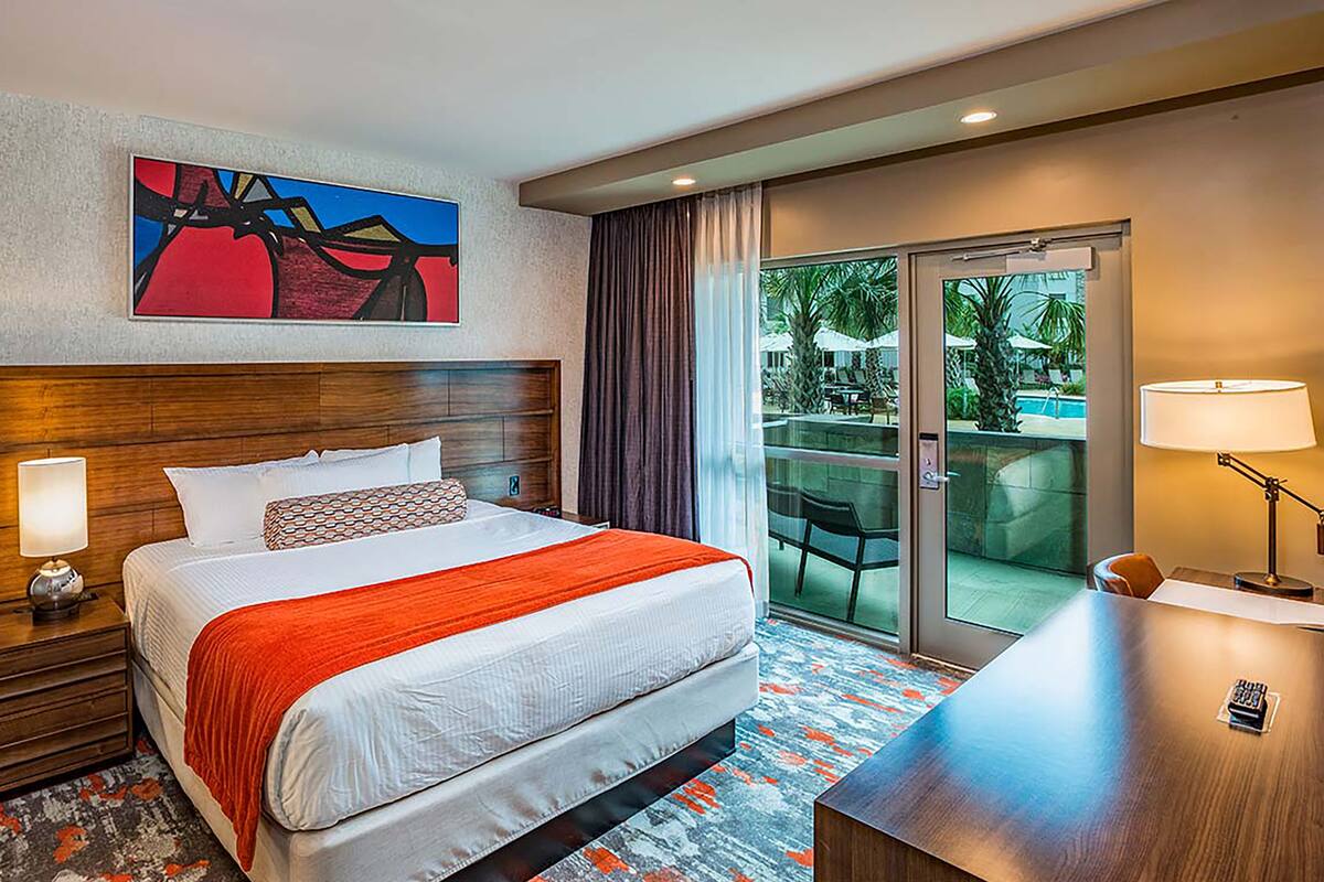 A hotel room with a bed, a desk, a chair, a nightstand, a painting, and a view of the outdoors.