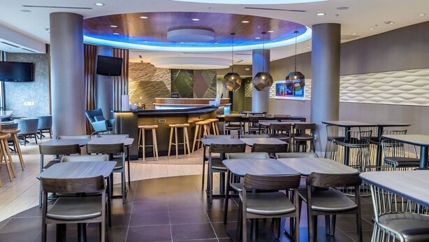 A modern restaurant with a curved counter, multiple seating areas, and a flat-screen TV mounted on the wall. - SpringHill Suites Houston NASA/Seabrook (Seabrook)