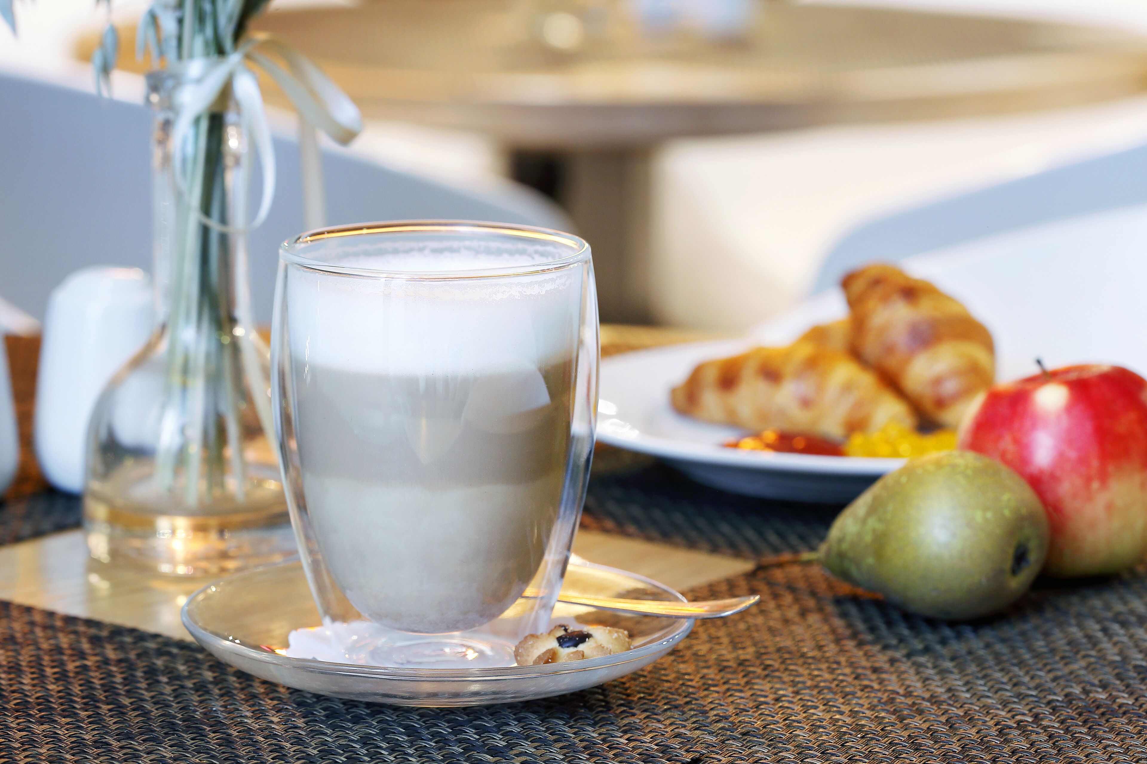 A glass of milk on a saucer, a plate with croissants and fruit, and a vase with dried flowers on a table.