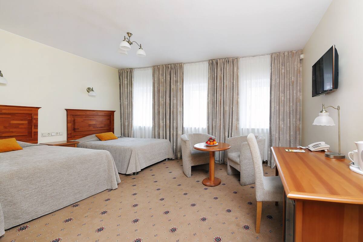 A hotel room with two beds, a desk, a chair, a table, a TV, and a lamp.