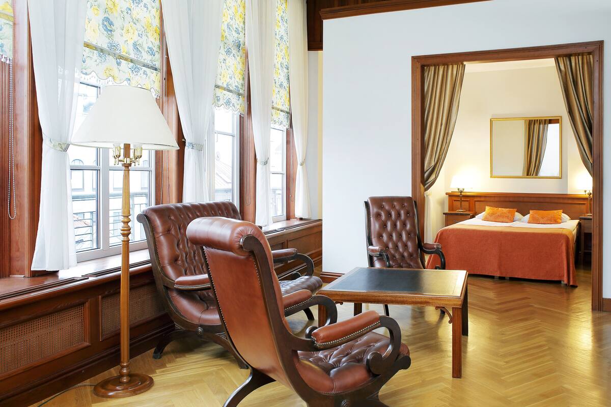 A hotel room with a wooden floor, a bed with an orange bedspread, a sitting area with leather chairs and a table, and a large window with curtains.