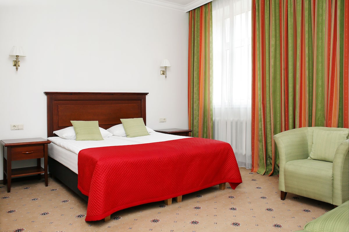 A hotel room with a bed, a nightstand, a green armchair, and a window with striped curtains.
