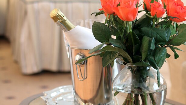 Una cama con ropa blanca y cabecera de madera, un jarrón con rosas anaranjadas, una botella de champán y un plato de fruta sobre una mesa de madera.