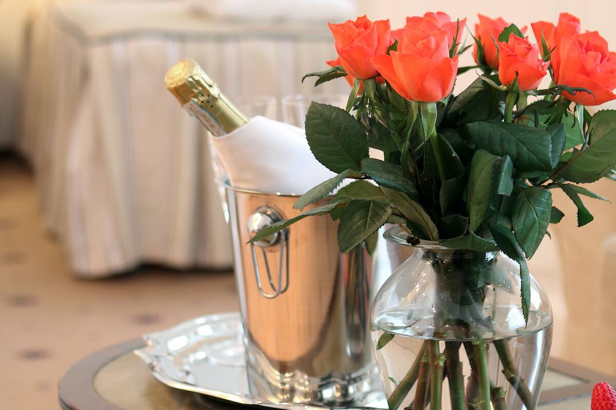 A bed with white bedding and a wooden headboard, a vase with orange roses, a champagne bottle, and a plate of fruit on a wooden table.