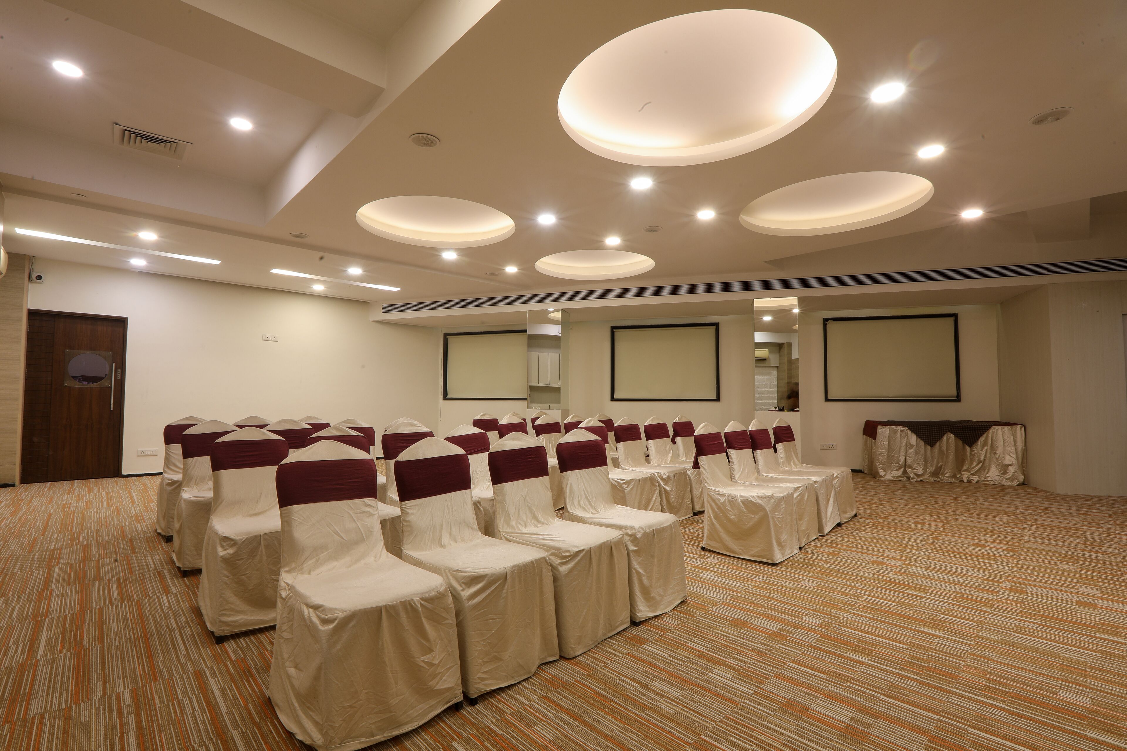Un salón de conferencias con filas de sillas cubiertas con manteles blancos y cintas granate, piso alfombrado e iluminación empotrada en el techo.