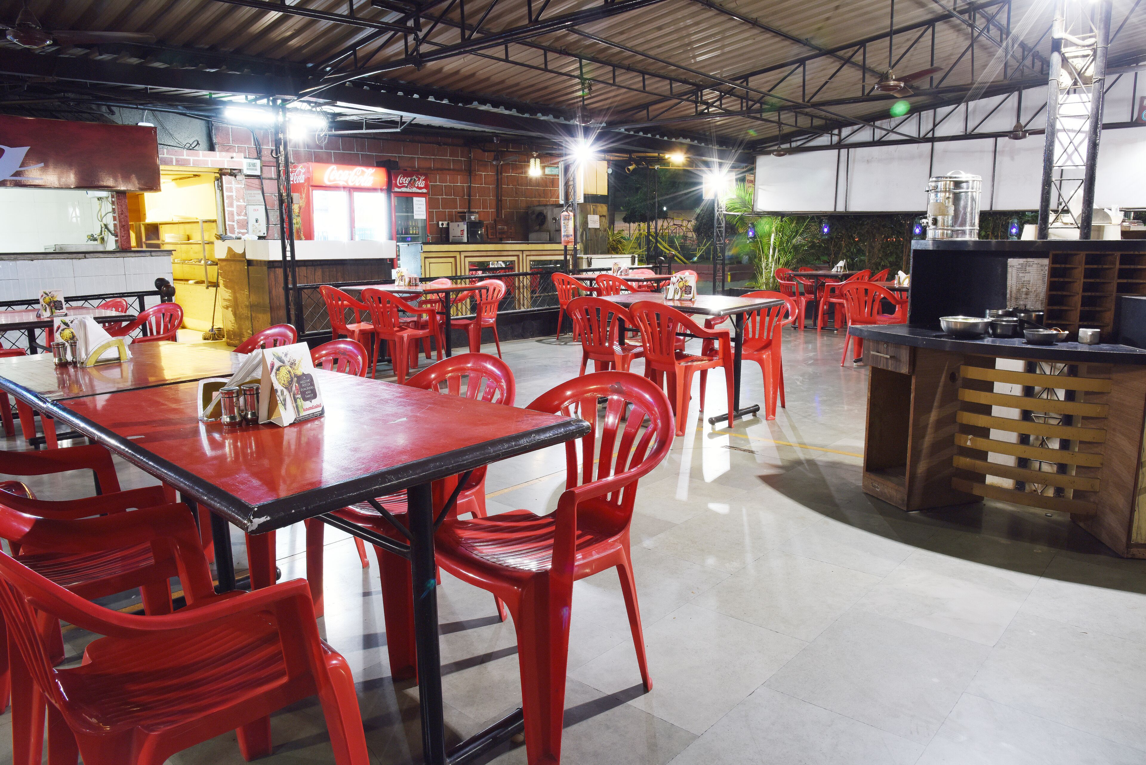 A spacious restaurant with red plastic chairs and tables, a Coca-Cola refrigerator, and a counter with kitchen equipment.