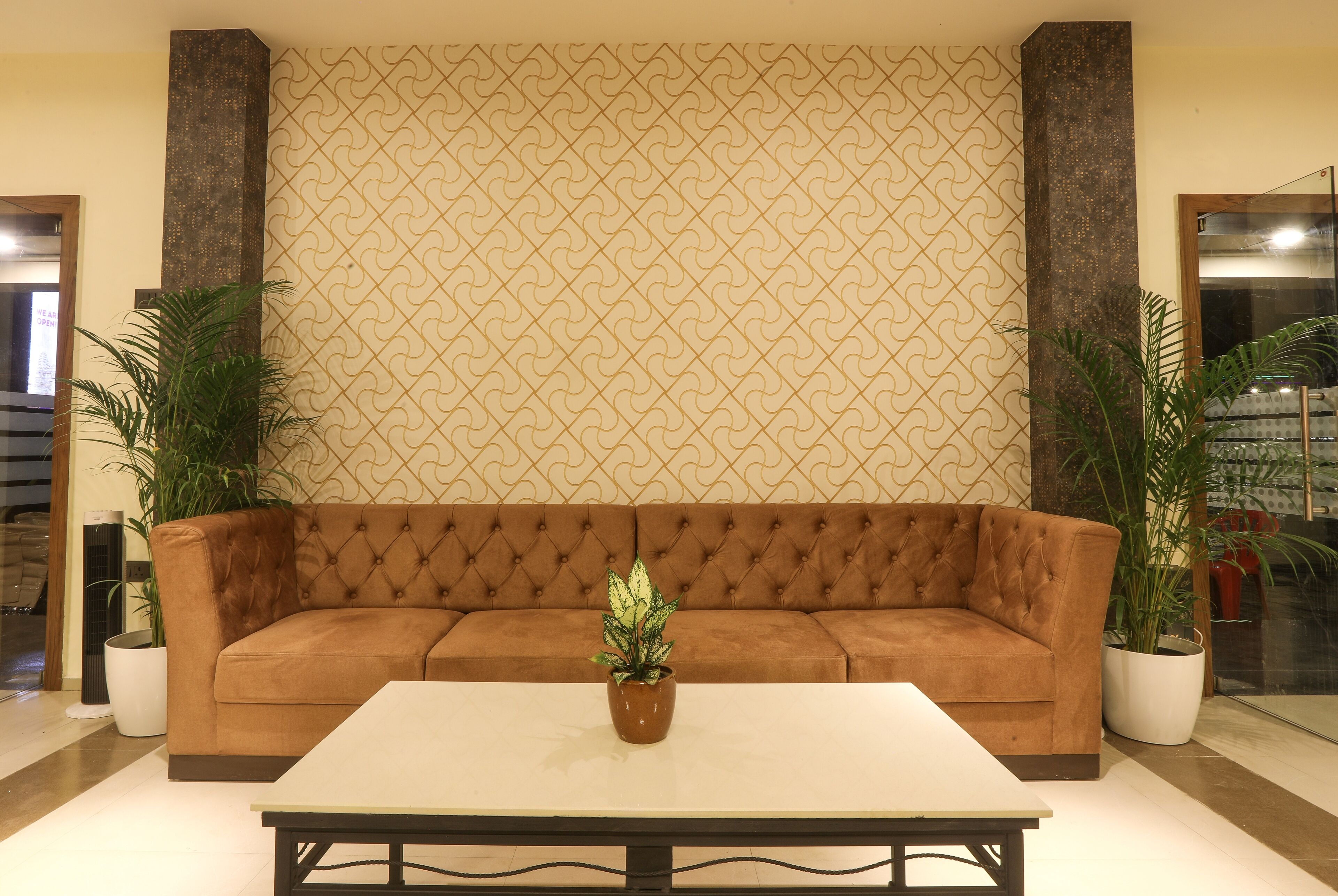 A brown sofa with a patterned wall behind it, a white table in front, and two potted plants on either side.