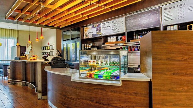 A modern coffee shop interior with a wooden counter, a refrigerated display case, and a menu board. - Courtyard by Marriott Biloxi North/D'Iberville (D'Iberville)