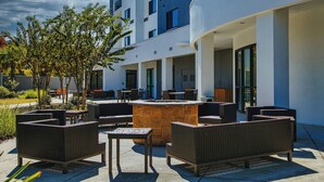 An outdoor seating area with wicker furniture and a central stone table, surrounded by a modern building with large windows and a curved facade. - Courtyard by Marriott Biloxi North/D'Iberville (D'Iberville)