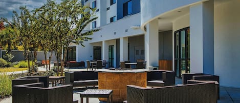 An outdoor seating area with wicker furniture and a central stone table, surrounded by a modern building with large windows and a curved facade.