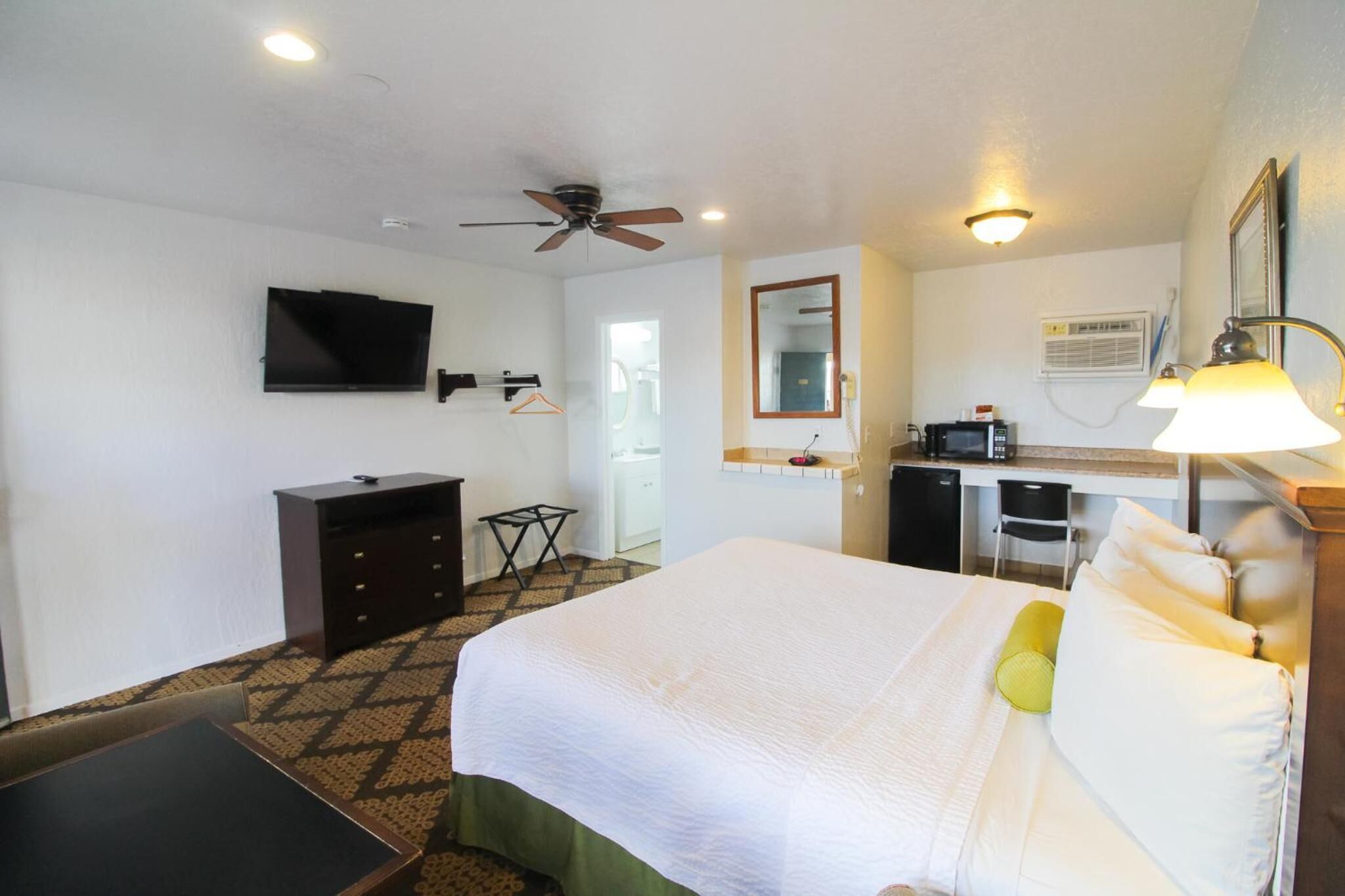 A hotel room with a bed, a desk, a television, a ceiling fan, and a kitchenette.