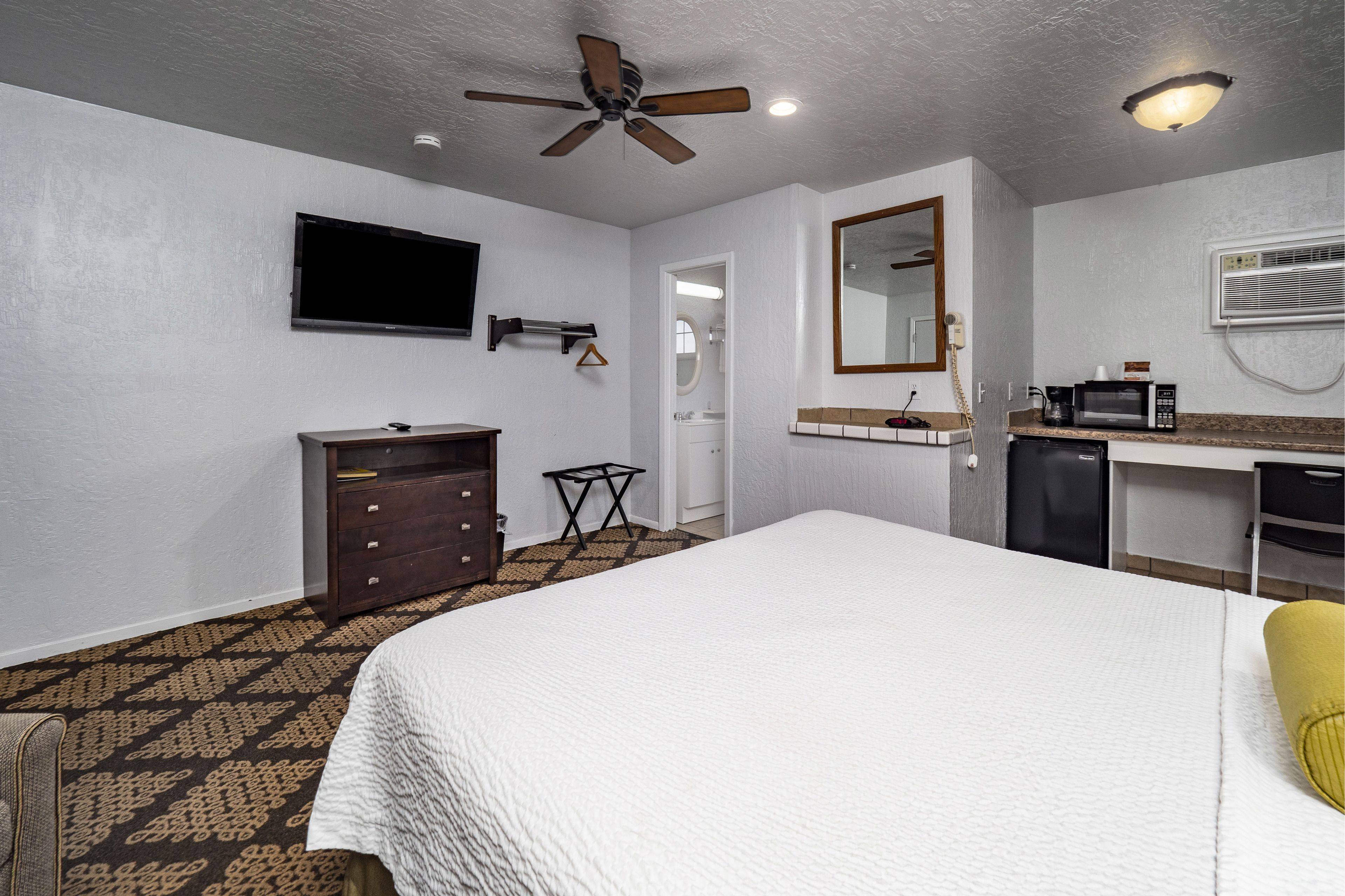 A hotel room with a bed, a desk, a microwave, a TV, a ceiling fan, and a bathroom visible through an open door.