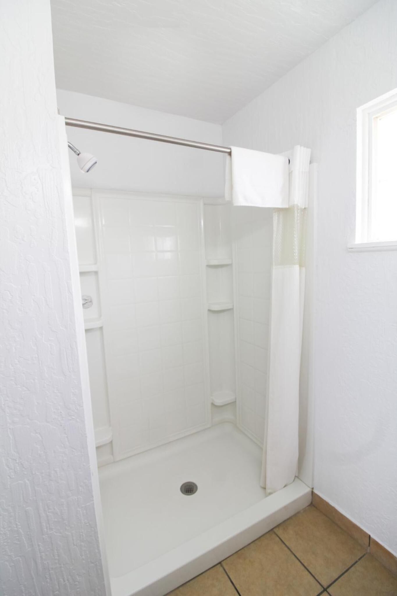 Un espacio de ducha pequeño y blanco, cerrado con cortina, con un perchero y una ventana.