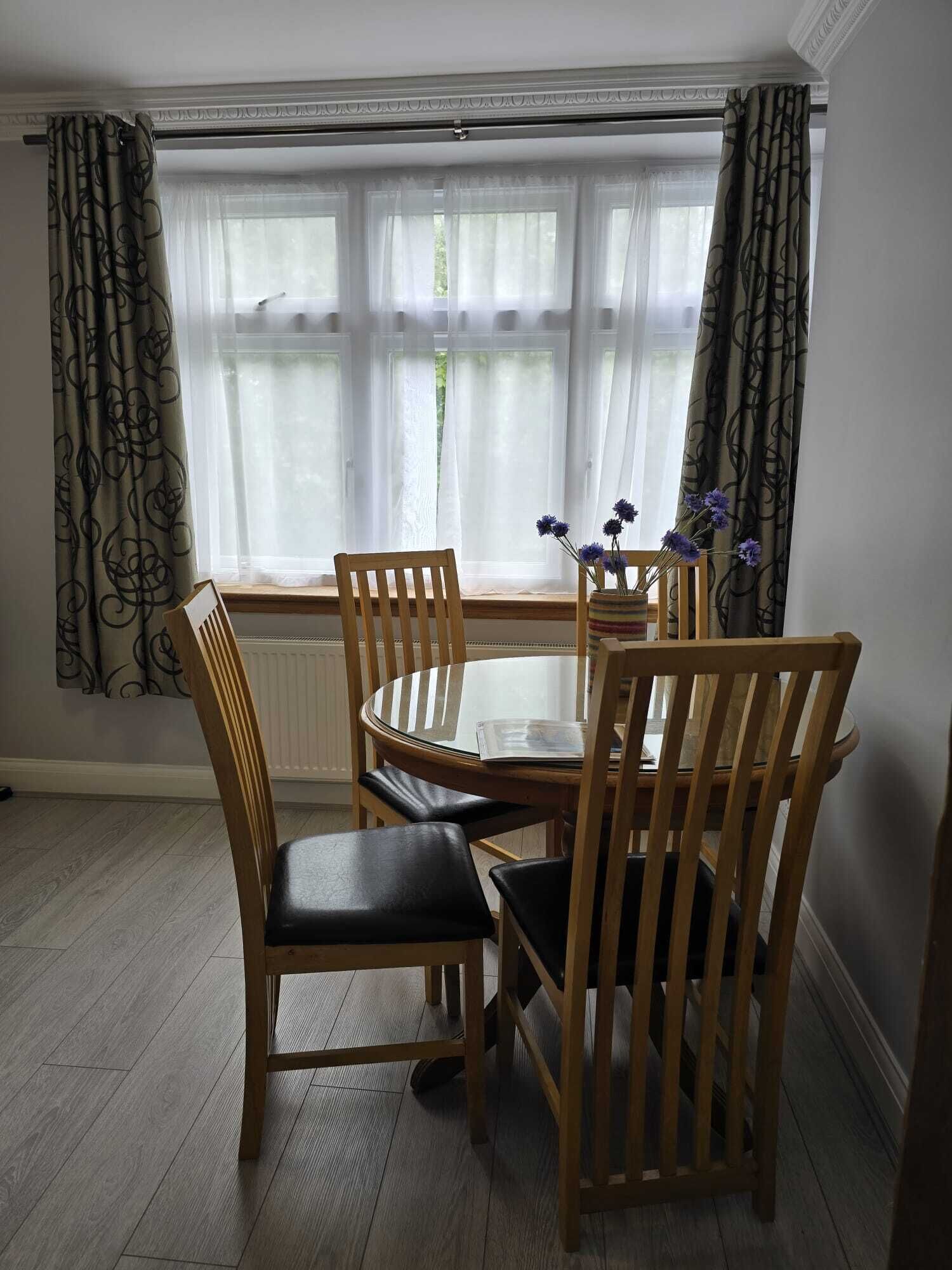 A dining area with a round table and four wooden chairs, a window with sheer curtains, and a vase with flowers on the windowsill.