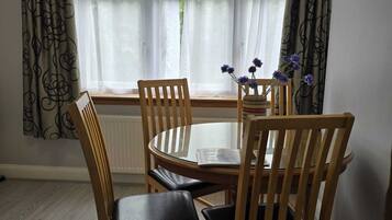A dining area with a round table and four wooden chairs, a window with sheer curtains, and a vase with flowers on the windowsill.