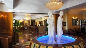 A lobby with a central fountain, marble statues, a reception desk, and a chandelier. - Aspen Apartments (London)