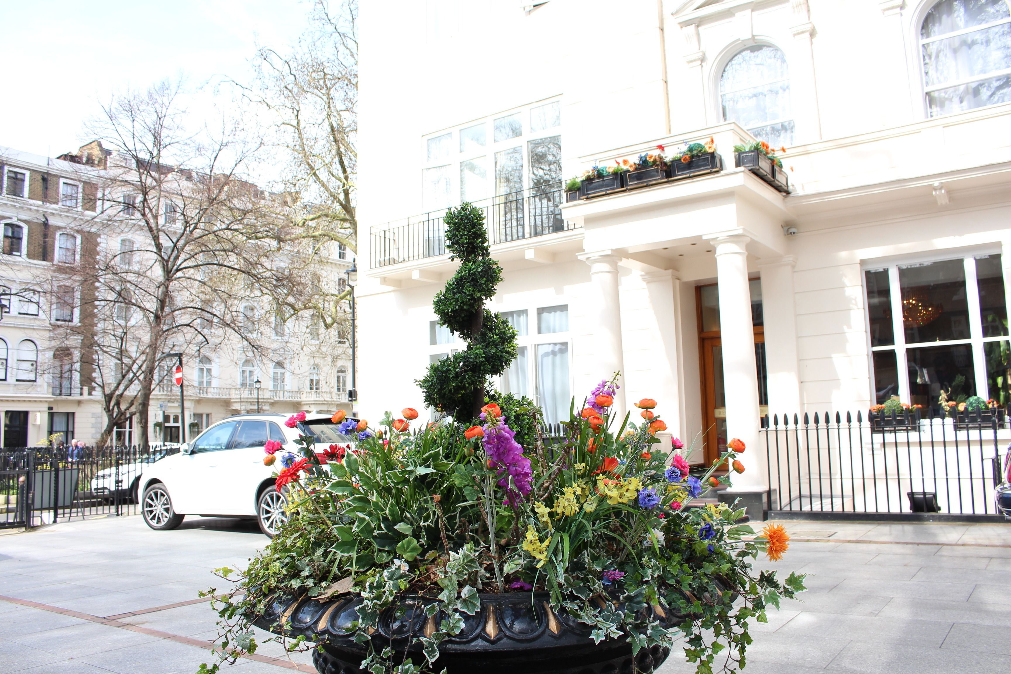 A white building with a black iron fence, a white car parked in front, and a flower bed with various plants and flowers.