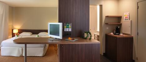 A hotel room with a bed, a desk with a computer, a small plant, and a refrigerator.
