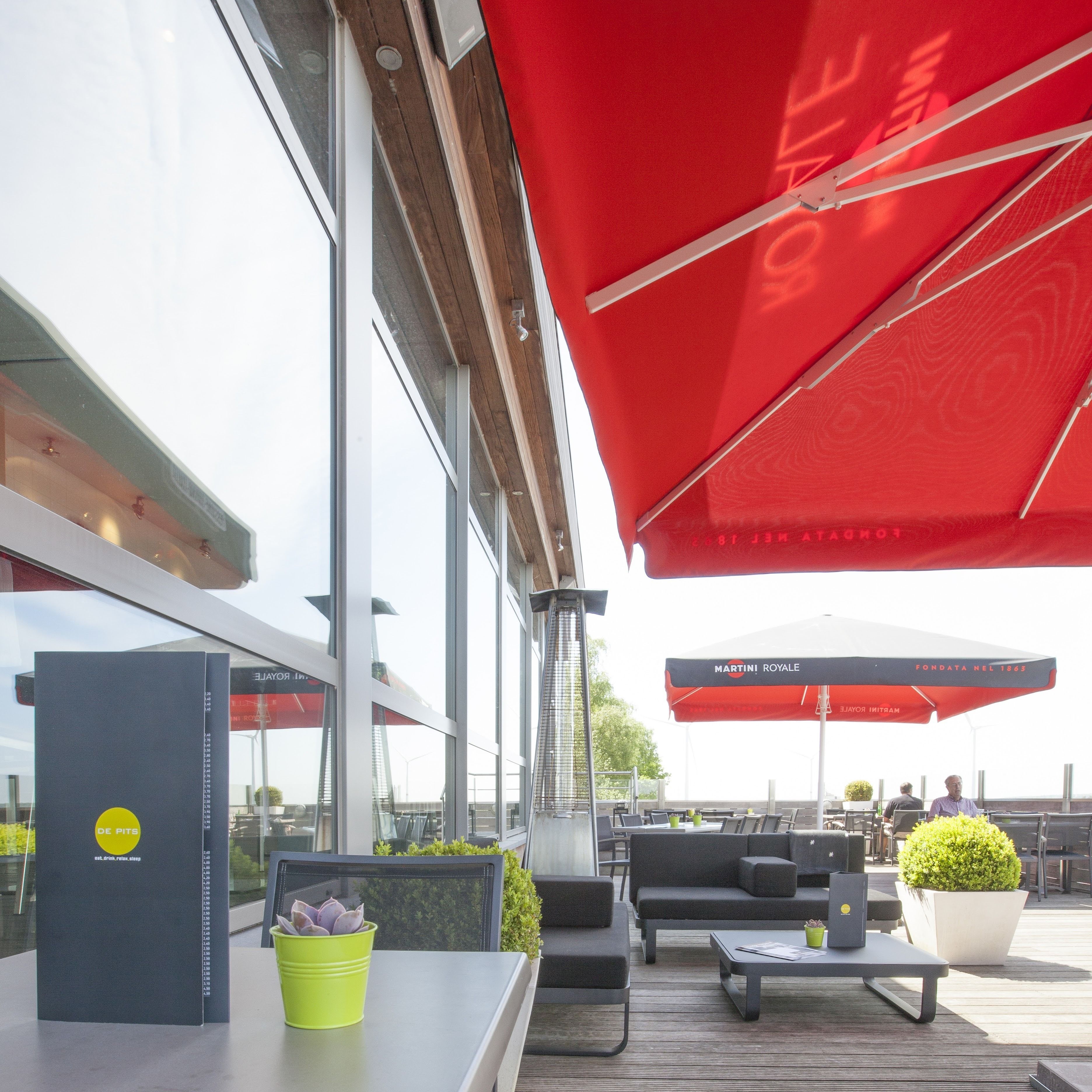 Un espace extérieur aménagé avec des chaises et des tables, des parasols rouges, une table sur laquelle se trouve un livre et une plante, et une vue sur la façade du bâtiment.