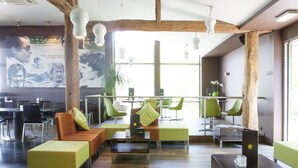 Een moderne zithoek met houten balken, groene en oranje zitplaatsen en een groot raam dat natuurlijk licht binnenlaat.