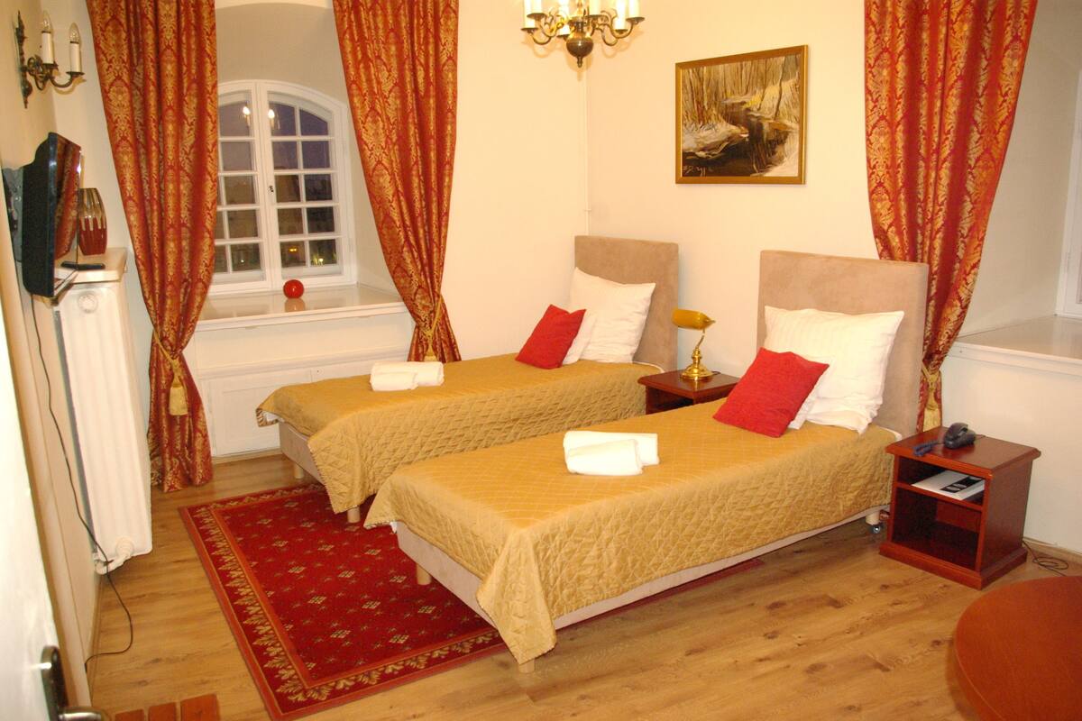 Een hotelkamer met twee bedden, een gordijn met een rood en goud patroon, een kroonluchter en een schilderij aan de muur.