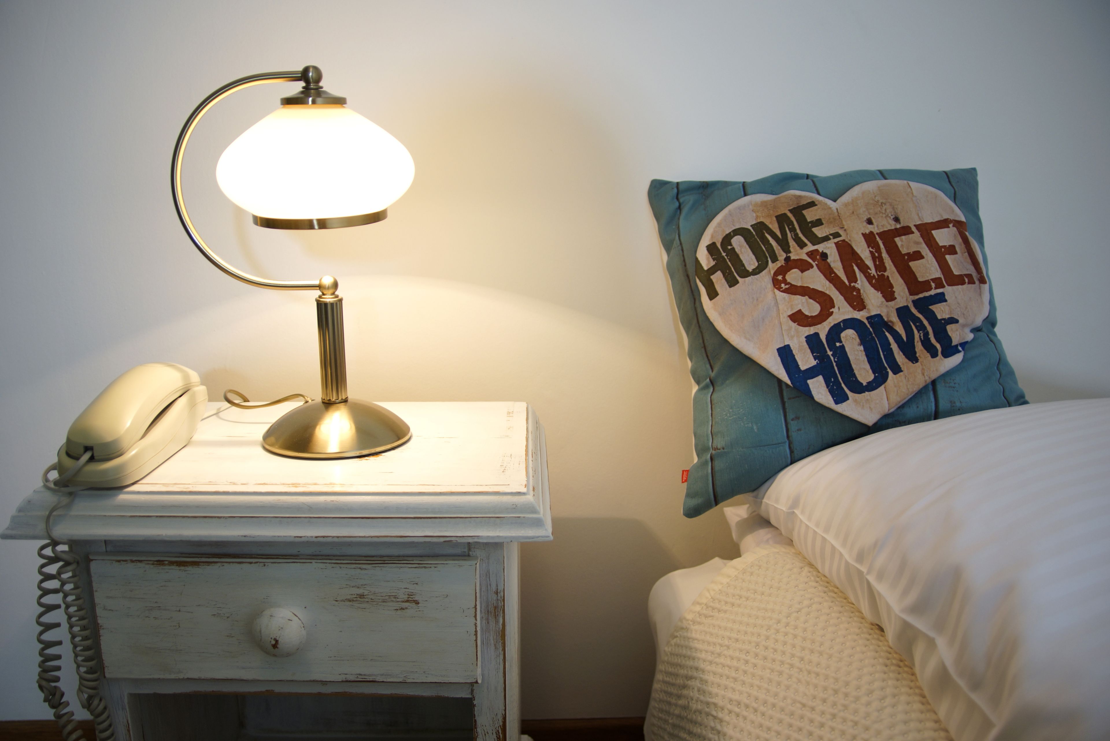 A bedside table with a lamp, a telephone, and a pillow with the text "HOME SWEET HOME".