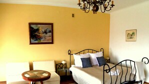 A bedroom with a bed, a chair, a small table, a chandelier, and two paintings on the wall.